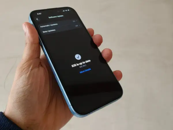 Dłoń trzyma iPhone'a z ekranem pokazującym "iOS is up to date iOS 26.2".