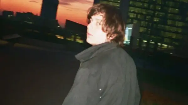 Francesco De Leo, con i capelli castani mossi, indossa un cappotto scuro. Sullo sfondo, un tramonto arancione illumina i profili degli edifici urbani.