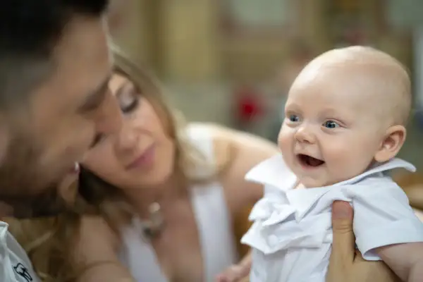 Un bébé souriant en tenue blanche, entouré de ses parents. Un moment précieux pour un baptême civil, depuis quand cette tradition existe-t-elle ?