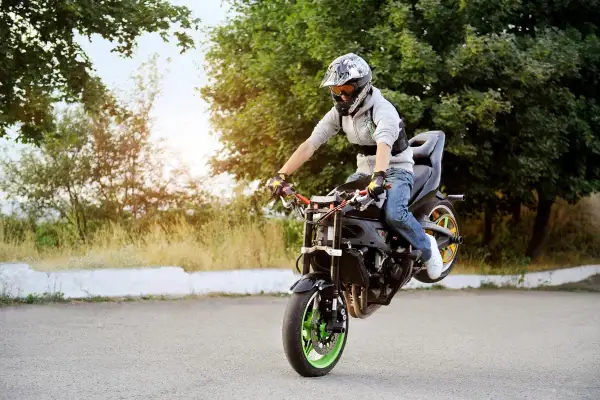 Motocyklista w kasku wykonuje jazdę na tylnym kole na asfalcie, otoczony zielenią.