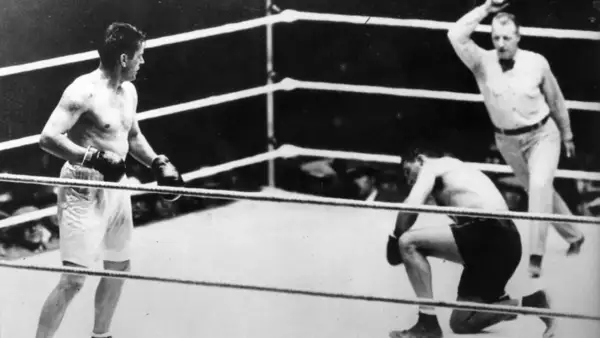 Jack Dempsey vs Gene Tunney in the ring. Dempsey stands, while Tunney is on one knee, with the referee looking on.