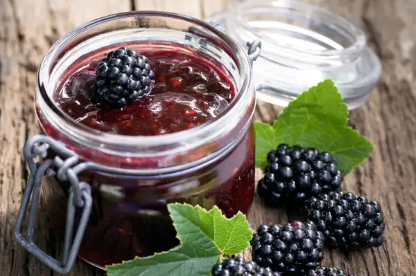 Glas mit Brombeermarmelade, daneben frische Brombeeren und Blätter. Perfekt, um sie mit Joghurt oder auf Toast zu mischen.