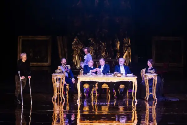 Scena z "Dziadów" w warszawskim teatrze. Przy złotym stole siedzą mężczyźni w strojach z epoki, obok kobieta na wózku.