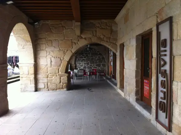 Arcadas de piedra en el casco antiguo de Galicia, con mesas y sillas de exterior. Los muros de piedra transmiten la historia de la zona.