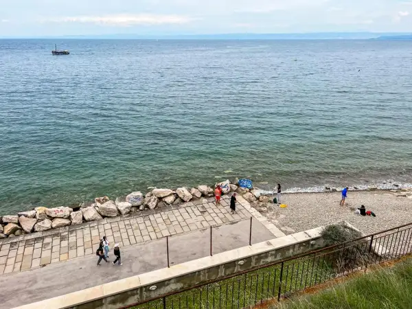 Spacerem po wybrzeżu w Słowenii Piran. Widać łódź na morzu i kilka osób odpoczywających na kamienistej plaży.