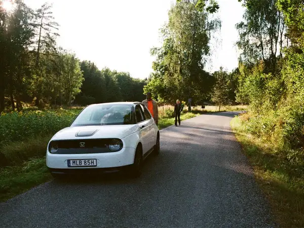 Vit elbil parkerad vid en skogsväg, redo för en bilsemester i Europa. Två personer står vid sidan av vägen.