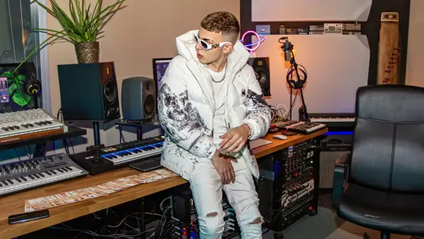 Un giovane skinny cantante con occhiali bianchi e giacca imbottita è seduto in uno studio di registrazione, circondato da tastiere e apparecchiature audio.