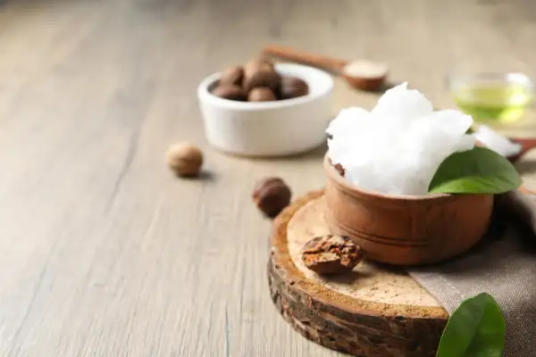 Un tazón de manteca de karité pura y cremosa, rodeado de nueces de karité y hojas verdes, sobre una superficie de madera rústica.