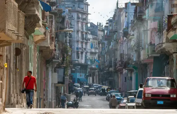 Photographie de rue d'une rue cubaine animée, avec des bâtiments colorés et délabrés, des voitures anciennes et un homme marchant seul.