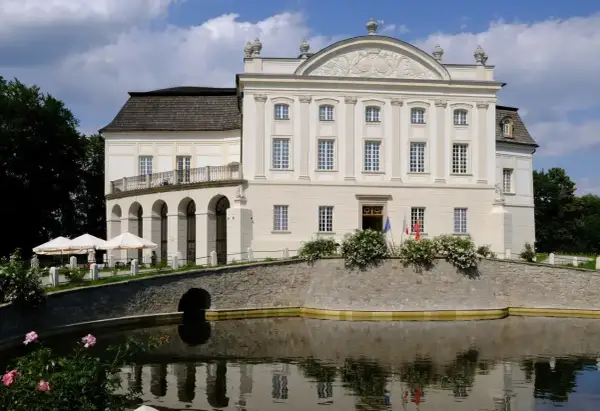 Pałac w Łącku, biała, okazała budowla z arkadowym tarasem, otoczona fosą i zielenią.