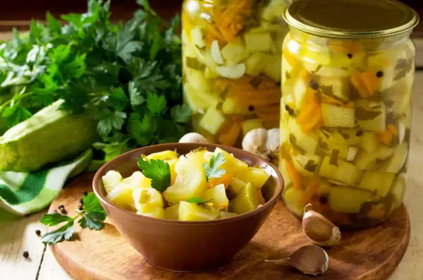 Słoiki z cukinią w zalewie słodko-kwaśnej z curry, miseczka z marynowaną cukinią, świeża cukinia, czosnek i natka pietruszki.