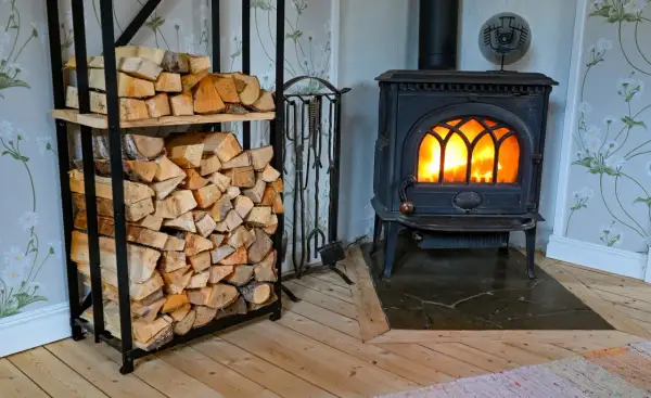 En mysig brasa sprakar i en gjutjärnskamin. Bredvid står en hylla fylld med ved, redo att installera braskamin för värme och trivsel.