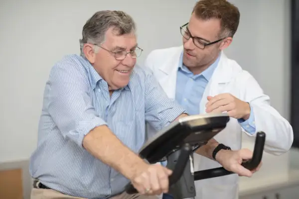 Un homme âgé sourit en effectuant un **test d'effort vélo**, supervisé par un médecin en blouse blanche.