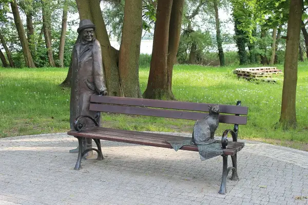 Pomnik mężczyzny w kapeluszu i kota na ławce w parku.