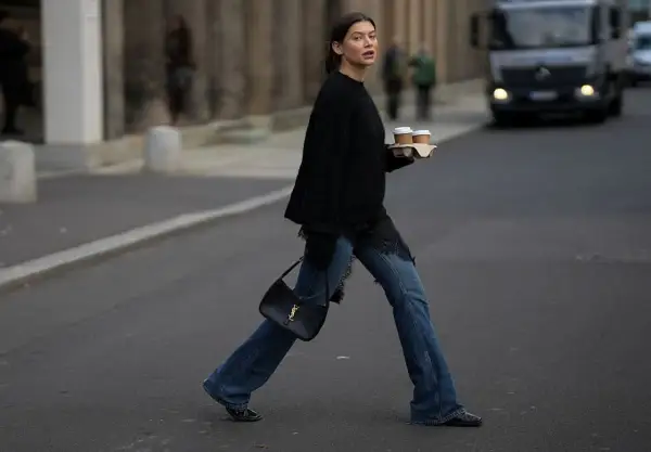 Kobieta w jeansach bootcut i czarnym swetrze z koronkowymi wstawkami niesie kawę. Stylowe jeansy bootcut to podstawa wielu casualowych stylizacji.
