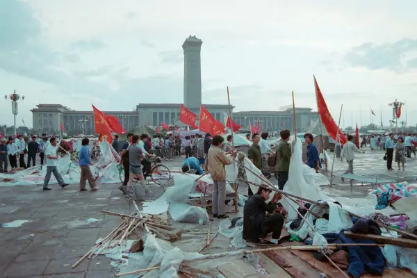 Ludzie na placu Tiananmen, czerwone flagi powiewają, budynki w tle, chaos i prowizoryczne namioty.