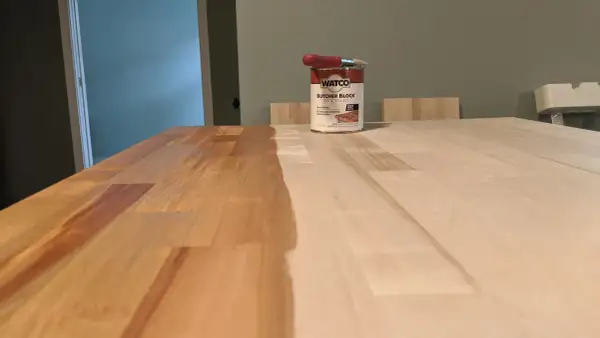 A can of Watco Butcher Block Oil & Finish sits on a wooden surface, with one half of the butcher block already stained and the other half awaiting treatment.