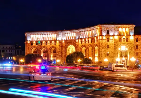 Nocna panorama placu w Erywaniu, największym mieście Armenii. Oświetlony budynek z arkadami, na ulicy widać ślady świateł samochodów i radiowóz.