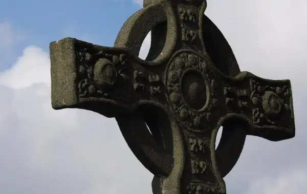 Kamienny krzyż celtycki z misternymi zdobieniami na tle błękitnego nieba z chmurami.