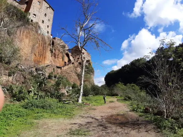 Sentiero nei pressi delle forre di Corchiano, con un edificio arroccato sulla rupe e un albero spoglio in primo piano.