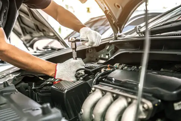 Mechanik w białych rękawiczkach dokręca śrubę kluczem nasadowym. Silnik samochodu jest otwarty, co sugeruje przegląd samochodu po terminie.