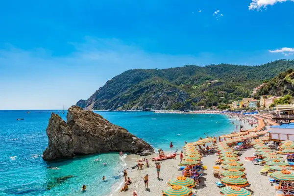 Spiaggia affollata sulla Riviera di Levante, con ombrelloni colorati, gente che nuota e montagne verdi sullo sfondo.