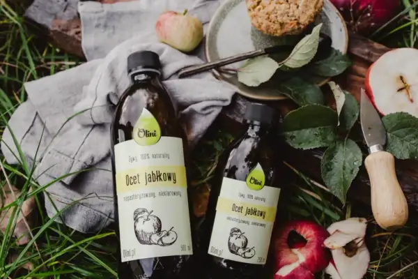 Dwie butelki naturalnego octu jabłkowego Olini, idealnego dla dzieci. Wokół jabłka i liście.