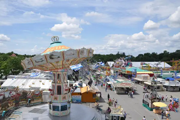 Buntes Treiben auf dem Volksfest Ingolstadt mit Kettenkarussell, Fahrgeschäften und Buden unter blauem Himmel mit Wolken.