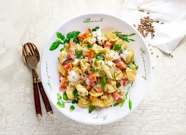 Pyszne tortellini z boczkiem i śmietanowym sosem, idealne jako pomysł na obiad. Posypane natką pietruszki.