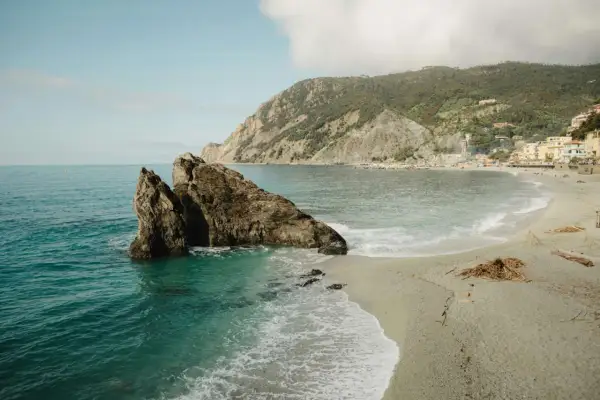 Un rocher émerge des eaux turquoise d'une crique. Une plage de galets s'étend vers un village coloré niché sur une colline verdoyante. Idéal pour savoir où partir en vacances cet été.