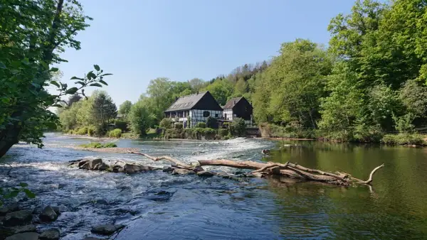Fachwerkhäuser am Flussufer, wo die **Wipperkotten wandern**. Ein Baumstamm liegt im rauschenden Wasser.