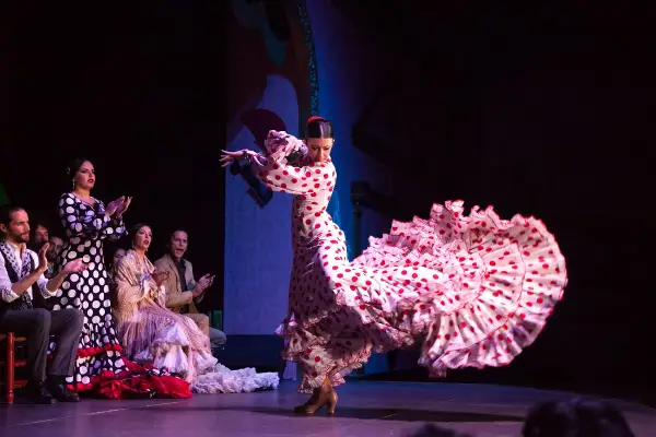 Bailarina de flamenco con un vestido de lunares rojos y blancos, su falda ondea con gracia. El público aplaude la seguidilla flamenca.