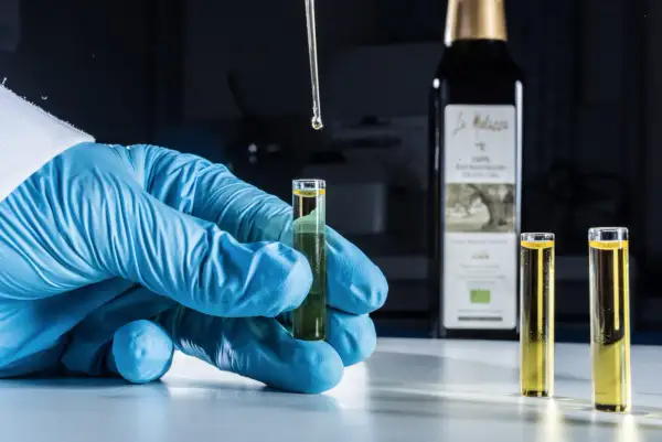 Analisi in laboratorio per verificare i perossidi nell'olio extravergine di oliva. Goccia di olio cade da pipetta in provetta tenuta da mano con guanto blu.