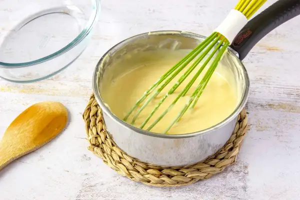 Un batidor verde mezcla una crema amarilla en una olla, mostrando cómo espesar un puré. Cerca hay una cuchara de madera y un recipiente de vidrio.