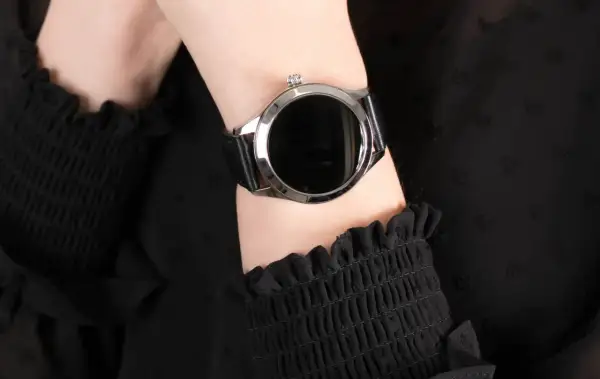 Damski smartwatch z czarnym paskiem na nadgarstku, na tle czarnej bluzki.