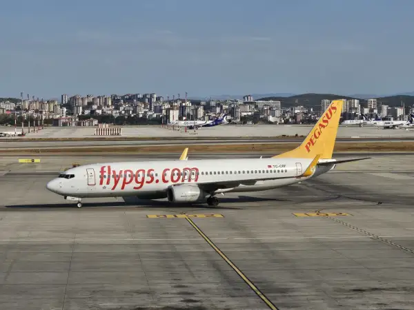 Samolot linii lotniczych Pegasus z napisem "flypgs.com" na kadłubie na płycie lotniska, w tle miasto.