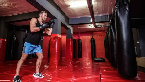 A boxer trains in a gym with many boxing bags. The red floor reflects the lights, and the boxing bag numbers are not visible.