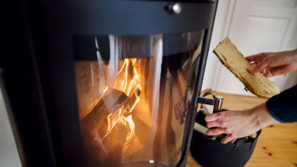 Eine Hand legt Holzscheite in einen brennenden Ofen. Die Flammen lodern im Inneren des Ofens.
