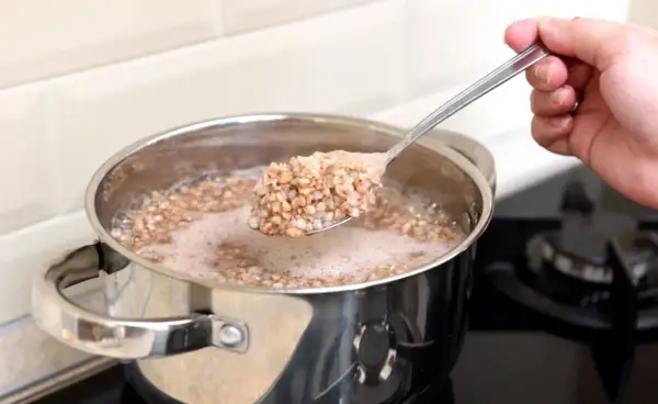 Eine Hand hebt einen Löffel mit dampfendem Buchweizen aus einem Topf, in dem Buchweizen gekocht wird.