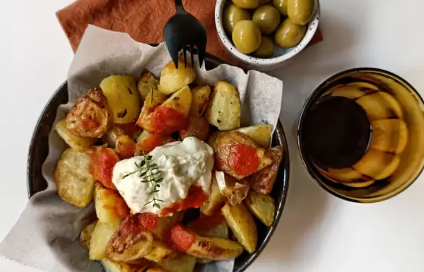 Patatas bravas con alioli, servidas con aceitunas y una bebida oscura.