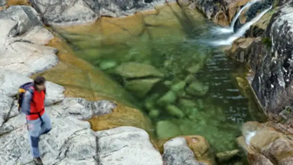 Senderista se detiene junto a un **río caldo** de aguas cristalinas y rocas musgosas.
