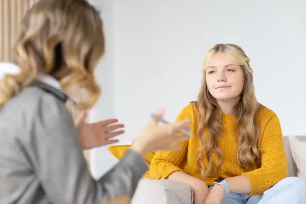 Nastolatka w żółtym swetrze podczas terapii. Rozmowa z terapeutką, która gestykuluje.