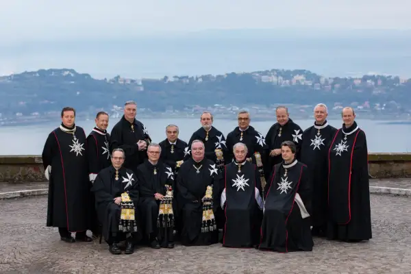 Uroczyste zgromadzenie członków zakonu maltańskiego w czarnych płaszczach z białymi krzyżami, na tle jeziora i miasta.