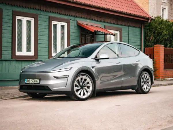Srebrny Tesla Juniper, nowoczesny samochód elektryczny, zaparkowany przed tradycyjnym, zielonym domem z czerwonym dachem.
