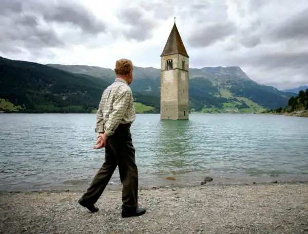 Ein Mann spaziert am Ufer eines Sees, auf dem ein Kirchturm aus dem Wasser ragt. Die Kirche im Stausee ist ein eindrucksvolles Zeugnis der Vergangenheit.