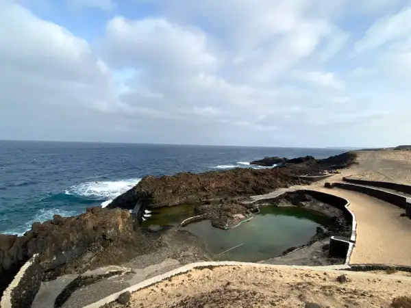 Piscinas naturales formadas por lava volcánica en la costa de Lanzarote, con el mar azul de fondo. No hay cocoteros, pero sí un paisaje salvaje.