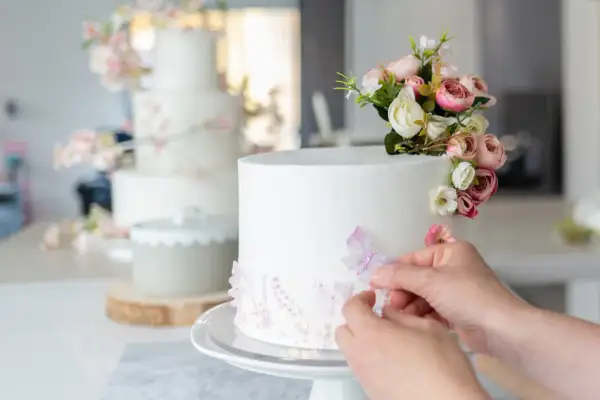 Una mano aggiunge una farfalla a una torta bianca decorata con fiori e paillettes. Sullo sfondo, un'altra torta a più piani con decorazioni floreali rosa.