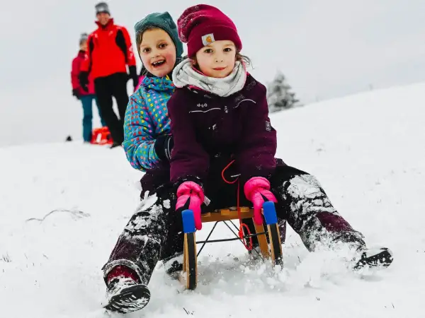 Zwei Kinder rodeln lachend einen verschneiten Hang im Bergischen Land hinunter.