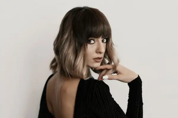 Kobieta z modnym, **blunt bob** w odcieniach brązu i blondu, w czarnej, eleganckiej sukience, z dłonią opartą o podbródek.