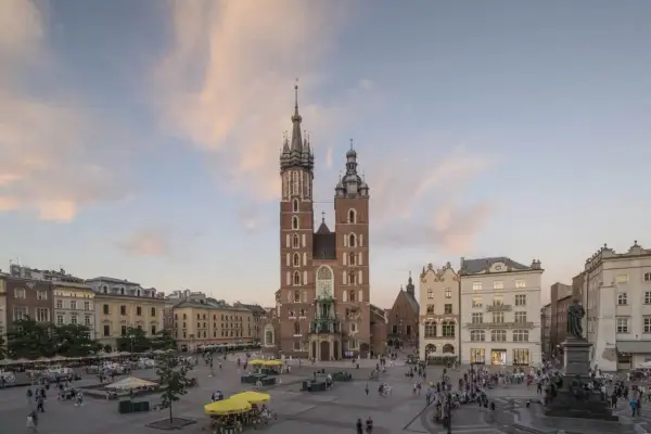 Krakowski Rynek Główny z majestatyczną Bazyliką Mariacką w tle. Ludzie spacerują, a nad nimi pastelowe chmury.